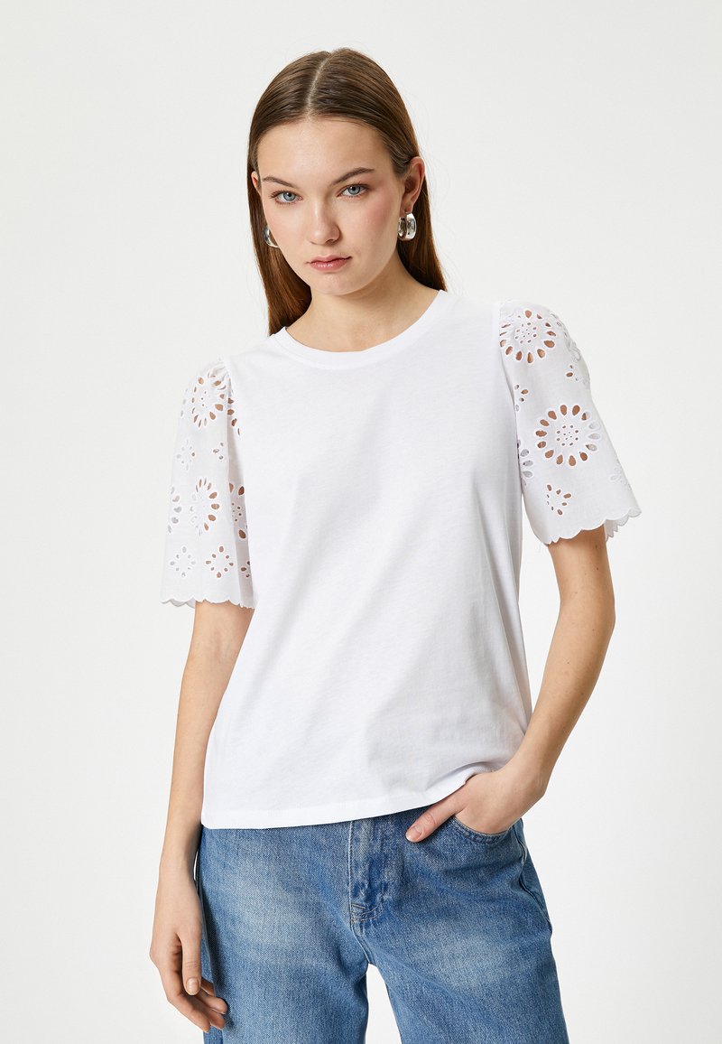 Hvid t-shirt med øjelet blonder kortærmer og bølgede kanter. Casual pasform med rund halsudskæring, parret med lyseblå denimjeans.