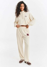 Krémově barevný fleecový pulovr s polovičním zipem a logem, doplněný o odpovídající tepláky s bordovou nášivkou a elastickým pasem.