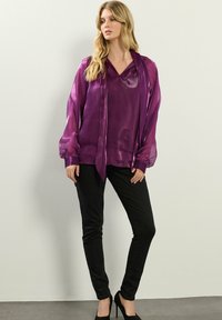Blusa viola semi-trasparente con scollatura a V e maniche lunghe a sbuffo, abbinata a jeans skinny neri e tacchi alti neri.