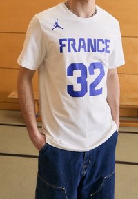 Biała bawełniana koszulka z niebieskim napisem "FRANCE" i numerem "32" z przodu. Na lewym ramieniu znajduje się małe niebieskie logo.