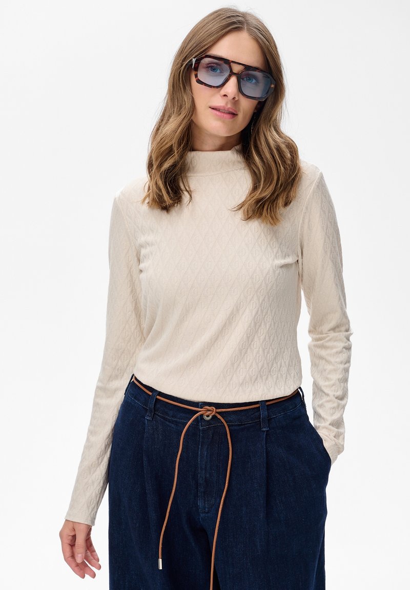 Vrouw met golvend haar draagt oversized schildpadbril, crèmekleurige textuurcol, donkerblauwe hoge taillebroek met een dunne bruine riem.