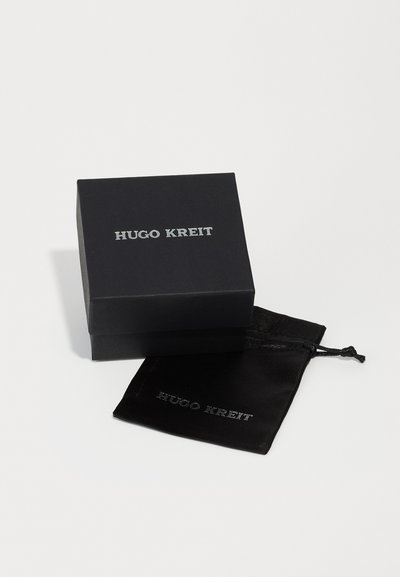Melnā kvadrātveida dāvanu kaste ar sudraba tekstu "HUGO KREIT" uz vāka, blakus atbilstošai melnai maisiņai ar to pašu zīmolu. Glīts, minimālisma dizains.