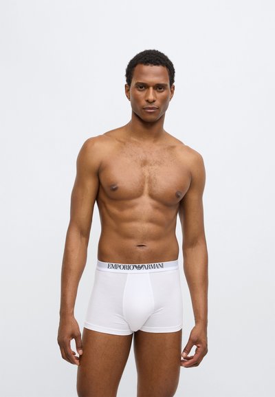 Boxer a costine in cotone bianco con taglio aderente, caratterizzati da una fascia elastica con logo in vita. Il modello ha un fisico tonico e sta in piedi in posizione eretta.