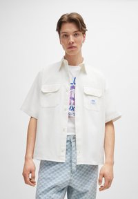 Weißes kurzärmliges Hemd mit zwei Brusttaschen, einem geraden Kragen und einer lockeren Passform, getragen über einem Grafik-T-Shirt und hellen karierten Jeans.