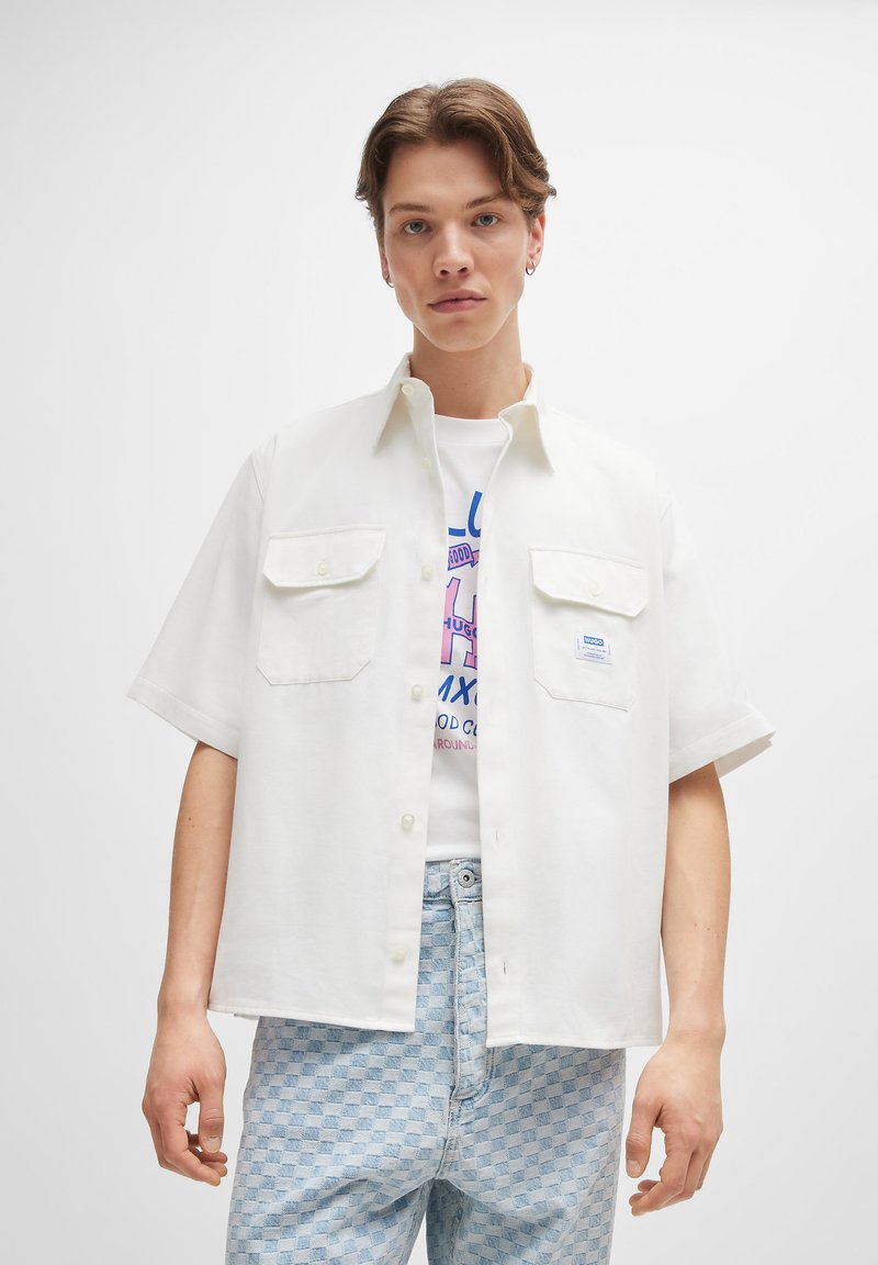 Weißes kurzärmliges Hemd mit zwei Brusttaschen, einem geraden Kragen und einer lockeren Passform, getragen über einem Grafik-T-Shirt und hellen karierten Jeans.