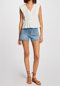 Top blanco sin mangas con escote en pico, detalles de botones y dobladillo peplum, combinado con pantalones cortos de mezclilla azul. El modelo lleva sandalias marrones con tiras.