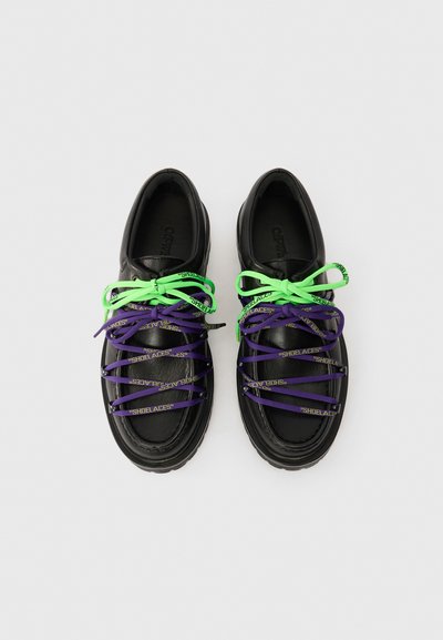 Μαύρα δερμάτινα παπούτσια με διπλές κορδόνες: φωτεινό πράσινο δεμένο στην κορυφή και μωβ με το κείμενο "ΚΟΡΔΟΝΑΚΙΑ" από κάτω, σε απλό ανοιχτό φόντο.