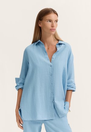 Woman with long straight hair wearing loose light blue button-up shirt and matching pants, hands in pockets, looking to the side against plain background.