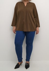 Brun, langærmet top med V-hals, båret med blå skinny jeans og sorte spidse sko. Glat tekstur og afslappet pasform.