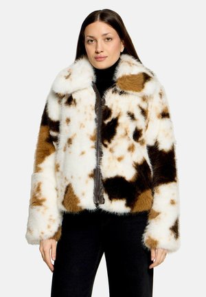 Femme portant une veste en fausse fourrure blanche, brune et noire à taches, par-dessus un col roulé noir et un pantalon noir, debout devant un fond uni.