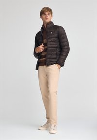 Puffjacka i mörkbrun, ribbad brun och tanrandig tröja, beige byxor och ljusa sneakers. Har en ståkrage och blixtlås framtill.