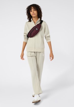 Beige sportjack en -broek, gecombineerd met witte sneakers. Rugzak is bordeauxrood met een ritsvak. Model heeft krullend haar.