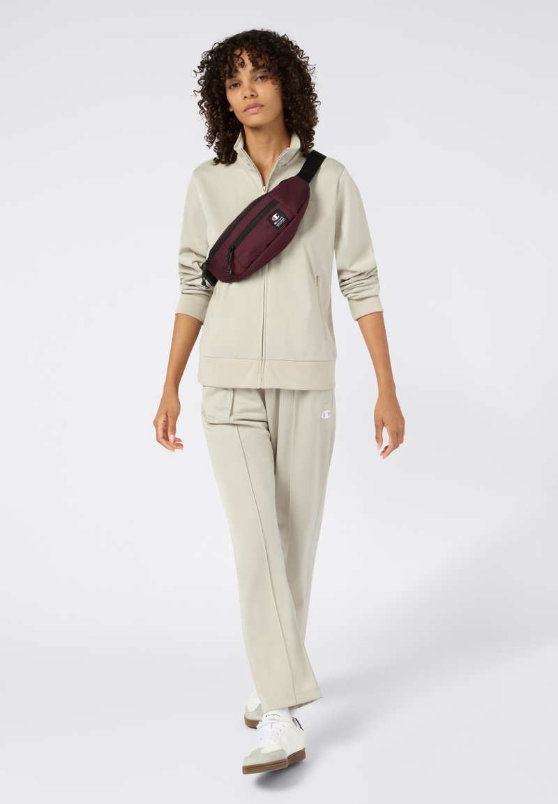 Giacca e pantaloni atletici beige, abbinati a sneakers bianche. Lo zaino è bordeaux con una tasca con zip. Il modello ha i capelli ricci.