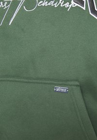 Zielona bluza z miękką fakturą, ozdobiona białym i czarnym haftem. Zawiera widoczną srebrną metkę marki oraz przednią kieszeń.