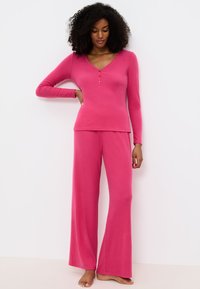 Roze tweedelige lounge set bestaande uit een lang-sleeve top met een V-hals en knoopdetail, gecombineerd met flared broeken. Zachte, textuurstof.