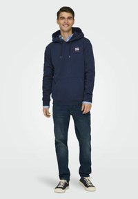 Dunkelblaues Hoodie mit Kordelzug, Fronttasche und Logo-Patch, kombiniert mit dunklen Jeans und Sneakern mit weißen Sohlen.