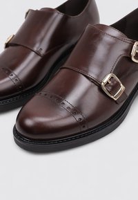 Zapatos de cuero marrón con un acabado pulido, que presentan un diseño de doble hebilla y hebillas doradas. La puntera tiene un detalle de brogue. Suela negra.