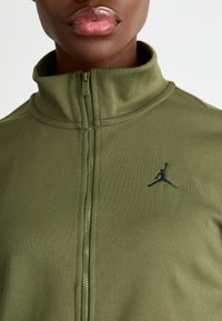 Oliven grønn jakke med glidelås, høy krage, teksturert stoff og en svart Jumpman-logo på venstre side.