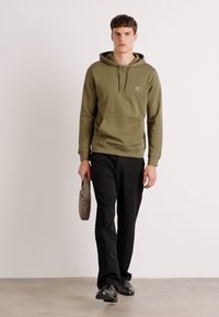 Olijfgroene hoodie met een voorzak, trekkoordcapuchon, gecombineerd met zwarte broeken en zwarte schoenen, met een grijze tas in de hand.