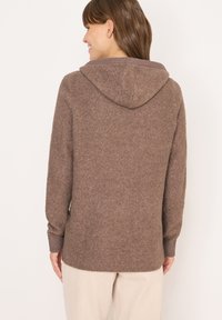 Brązowy sweter z dzianiny z kapturem, z prążkowanymi mankietami i dołem. Miękka tekstura i luźny krój, widoczne detale szwów.