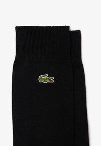 Calcetines de algodón negros con textura acanalada, que presentan un logo de cocodrilo bordado en verde. El diseño incluye un puño alto para mayor comodidad.