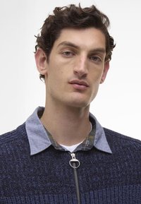 Marineblauwe zip-up trui met een getextureerd breipatroon en lichtgrijze kraag over een geruite shirt. Close-up weergave richt zich op de details van de kraag en de rits.