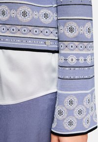 Knit sweater with horizontal purple and black stripes, featuring circular patterns in cream, blue, and metallic threads; smooth white underlayer.