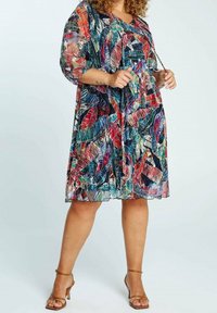 Nainen, jolla on päällään polvipituinen, värikäs lehtikuvioinen mekko, jossa on läpinäkyvät hihat, sekä ruskeat nauhakitkakuulahihtokengät yksinkertaista taustaa vasten.