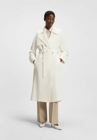 Weißer Trenchcoat mit breiten Revers, Gürtel und Seitentaschen. Getragen über beigefarbenen, weit geschnittenen Hosen und passenden Absatzschuhen. Glatte Stoffstruktur.