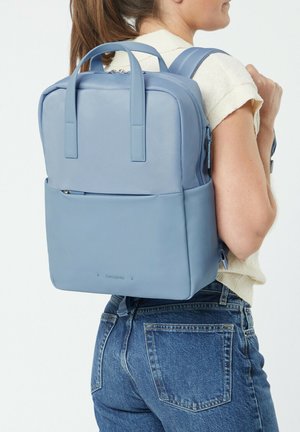 Hellblauer Rucksack aus glattem Material mit strukturierter Form, zwei Griffen und einer Reißverschlusstasche vorne.