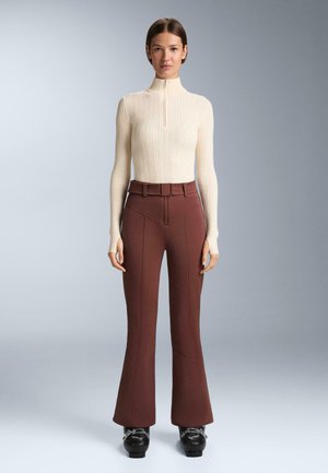 Cremfarbener, gerippter Rollkragenpullover mit kurzem Reißverschluss und langen Ärmeln, kombiniert mit bordeauxfarbenen, ausgestellten Hosen mit hervorgehobenen Nähten und einem Gürtel. Schwarze Schuhe.