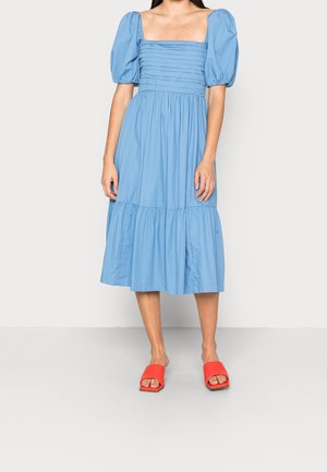 Mujer con vestido midi azul claro con mangas abullonadas y escote cuadrado, combinado con zapatos rojos abiertos y sin tacón.