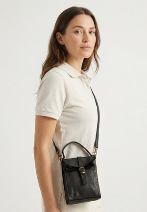 Vrouw draagt een beige poloshirt met een zwarte leren crossbodytas met een gesp aan de voorkant en een bovenste handvat.