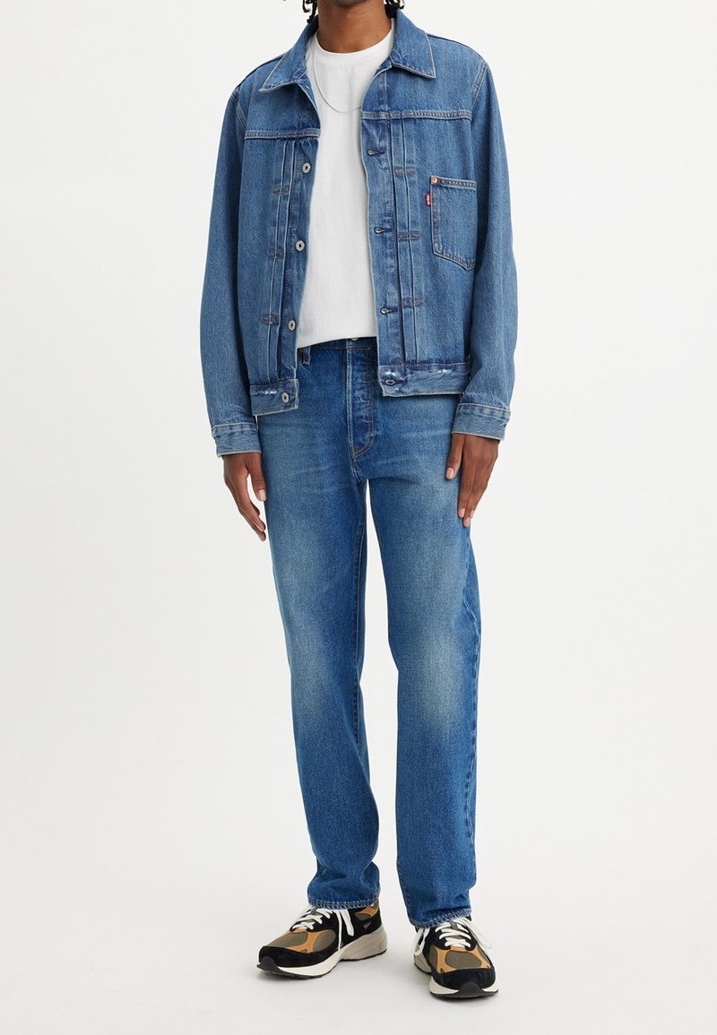 Homme portant une veste en jean bleu, un t-shirt blanc, un jean bleu et des baskets noires, camel et blanches, debout devant un fond uni.