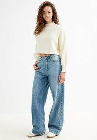 Suéter corto de punto acanalado crema combinado con unos pantalones vaqueros de tiro alto, de corte holgado y en azul claro, y zapatillas blancas.