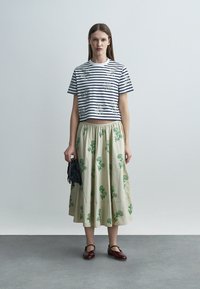 Nő csíkos, virágos hímzésű crop topot, bézs szoknyát zöld virágmintákkal, bordó Mary Jane cipőt visel, és egy kis fekete bojtos kézitáskát tart a kezében.