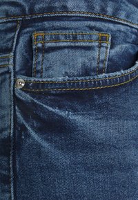 Jeans de ganga azul escuro com um bolso frontal apresentando costura amarela e um botão prateado. O tecido mostra um ligeiro desgaste e variações na textura.