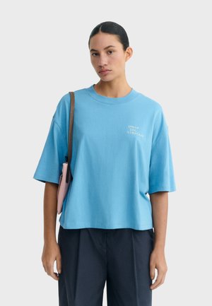 Kobieta o ciemnych włosach w niebieskiej, oversize'owej koszulce z małym białym napisem na piersi, czarnych spodniach oraz z różowym paskiem torby na ramieniu.