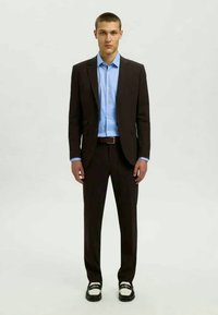 Costume marron avec une coupe ajustée, chemise bleu clair en dessous, ceinture en cuir, associé à des chaussures noir et blanc, sur fond neutre.