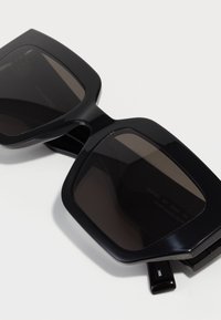 Lunettes de soleil noires surdimensionnées avec des verres carrés. Fabriquées en matériau glossy, avec une teinte foncée. Les détails incluent une étiquette en résine d'origine végétale et une charnière.