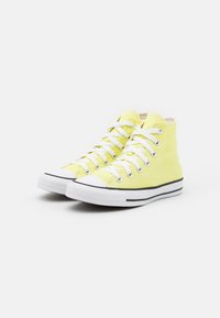 Pár žlutých vysokých plátěných tenisek s bílými gumovými podrážkami a bílými tkaničkami na bílém pozadí.