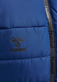 Blå vadderad jacka med ett strukturerat rutmönster, som har en svart "hummel"-logotyp och kontrasterande svarta dragkedjedetaljer.