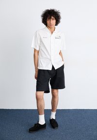 Short-sleeve white button-up shirt, black shorts, black shoes, and white socks, featuring an embroidered pocket detail and a casual fit.