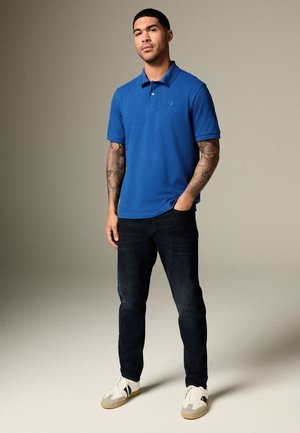 Mees, kellel on tätoveeritud käed, kannab sinist polosärki, musti teksaseid ja valgeid tenniseid, seisab ühe käega taskus tavalise tausta ees.