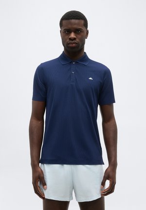 Mees seisab näoga ettepoole, kandes tumesinist polo särki ja heledaid halli värvi pükse, ilma mustrita taustal.