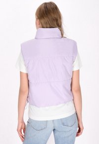 Gilet imbottito senza maniche di colore viola chiaro, con colletto, caratterizzato da cuciture orizzontali e cordoncino regolabile all'orlo, indossato sopra una camicia bianca.