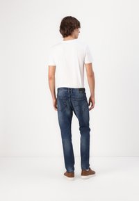 Jeans de mezclilla azul con un corte ajustado, costuras visibles y dos bolsillos traseros, combinados con una camiseta blanca lisa y zapatillas marrones.