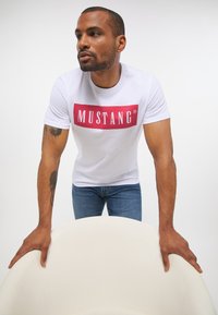 Biała bawełniana koszulka z dużym czerwonym prostokątnym logo "MUSTANG". Model ma na sobie niebieskie dżinsy i opiera się o białe, zakrzywione krzesło.