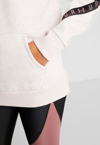 Personne portant un sweat-shirt rose clair avec le logo Under Armour sur la manche et des leggings à blocs de couleurs noir et rose, la main dans la poche de la capuche.