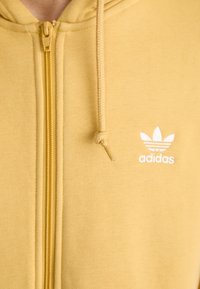 Žlutá mikina na zip z měkké látky, s bílým logem Adidas a strukturovaným vzorem podél zipu. Kápě má šňůrku na stažení.